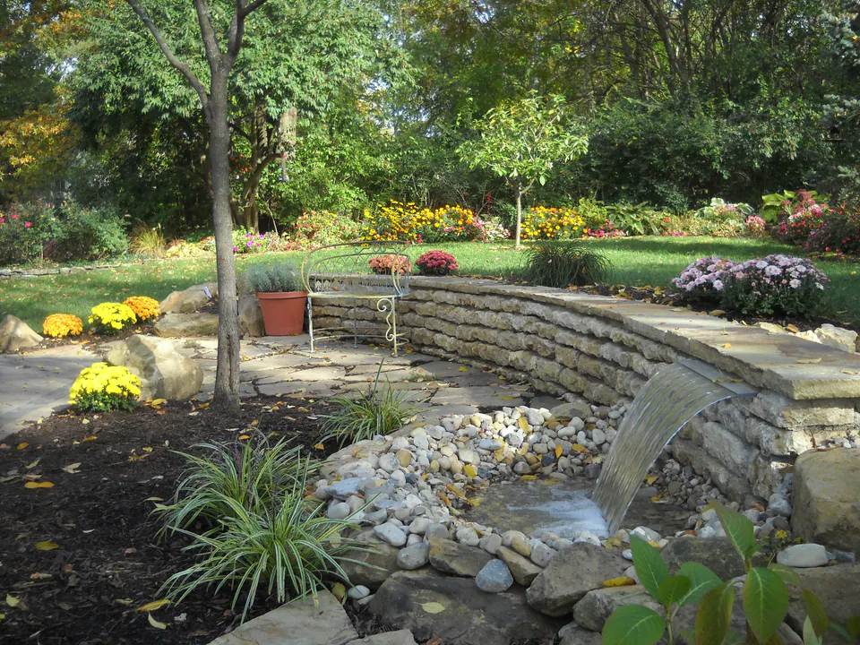 Water spilling from a curved stone wall into a rock-lined stream bed with fall flowers